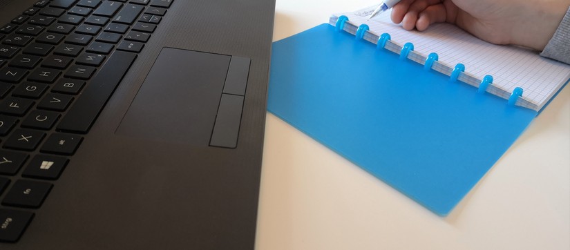 Neben der Tastatur eines Laptops macht eine Person in einem blauen Notizbuch Notizen zur Aufnahme.